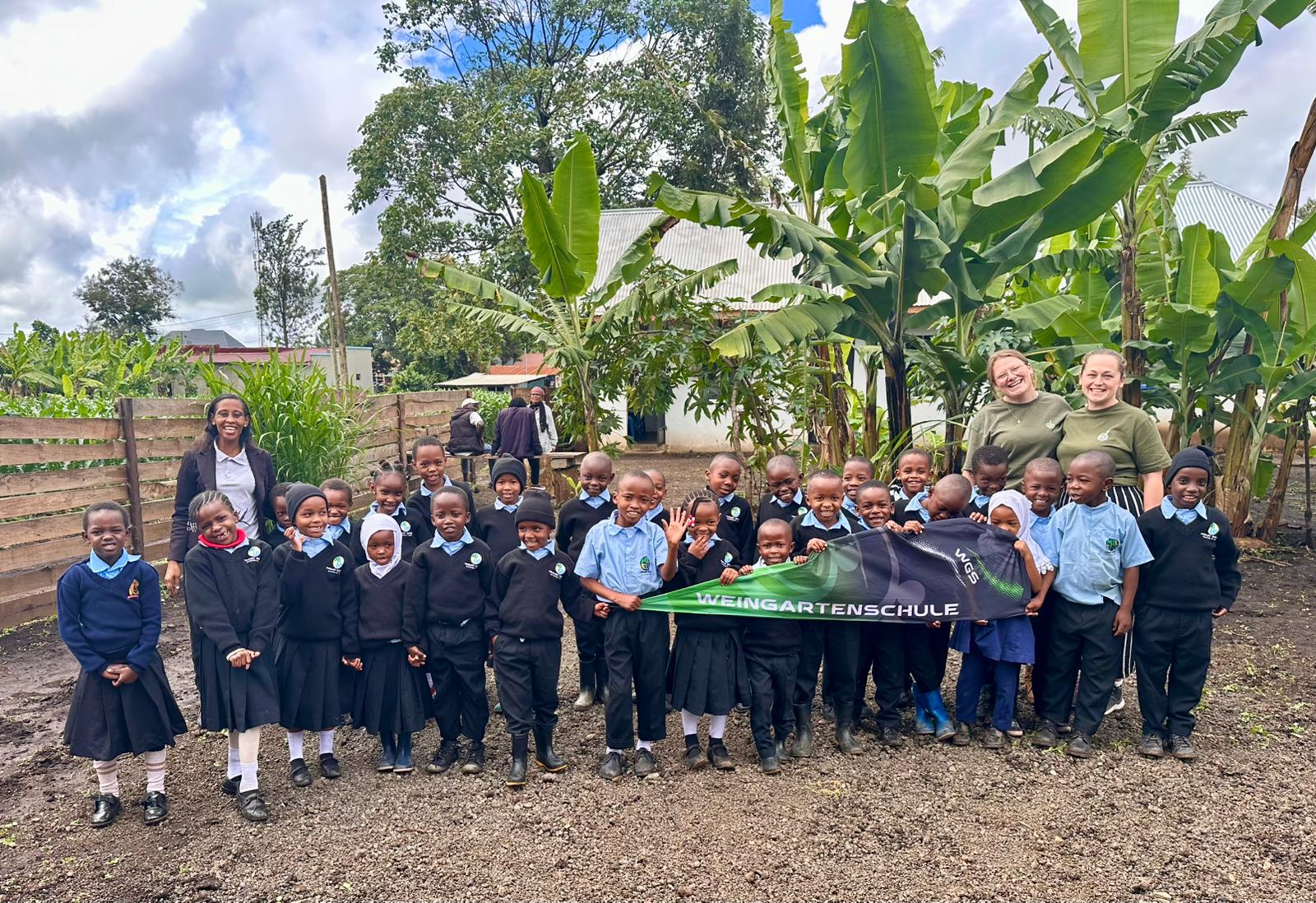 Die Kinder der MILELO Primary School in Schuluniform stehen mit zwei Lehrkräften der Weingartenschule vor Bananenstauden und halten ein Banner der Weingartenschule